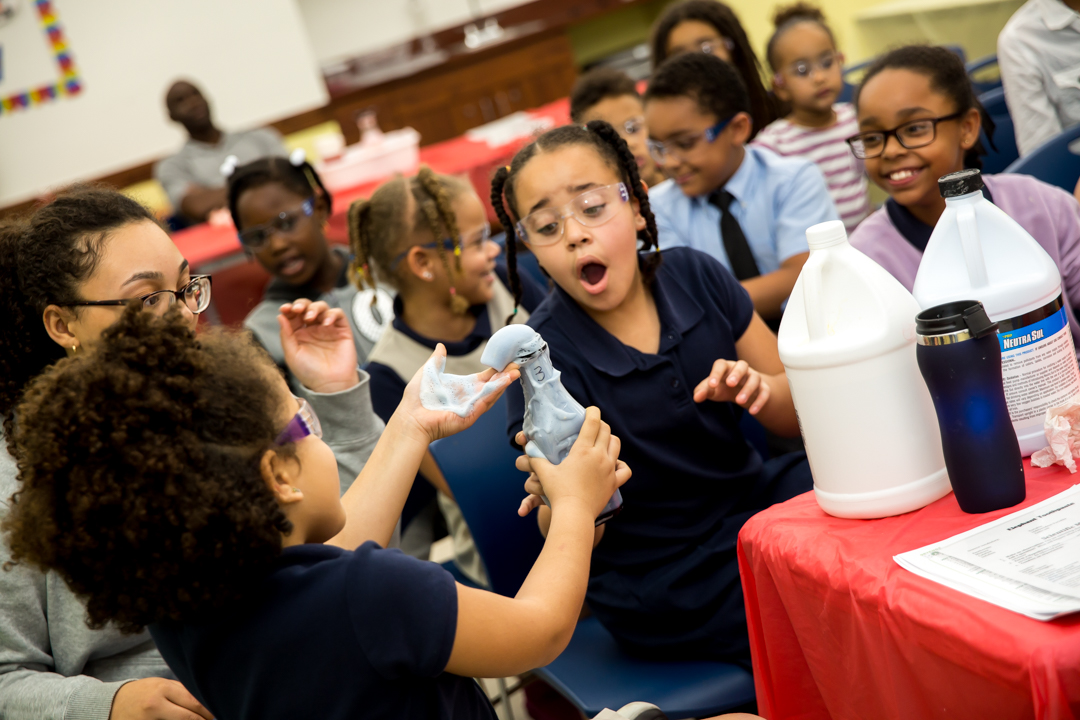 Children wearing goggles watch as a water bottle fills with foam during a science experiment.
